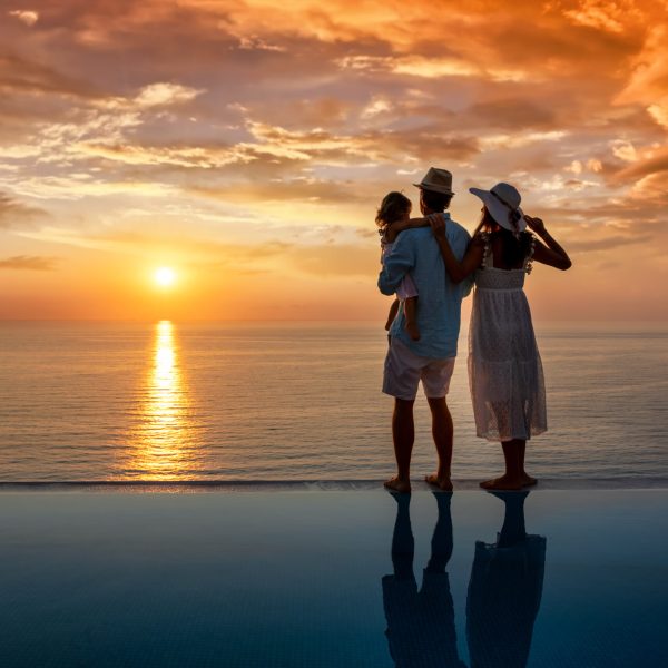A family on summer vacation standing by the swimming pool enjoys the view to the romantic sunset over the sea